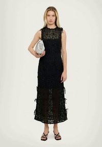 Svart blondekjole med solid skjørt, ermeløs design, dekorative bånd på sidene og en strukturert overflate. Modell som holder en sølvfarget clutch.