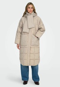 Beige lång vadderad kappa med hög krage och matchande scarf. Har två framfickor och rak skärning, kombinerad med blå vida jeans.