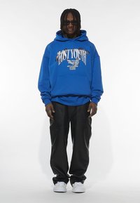 Felpa blu oversize con stampa grafica, tasca a marsupio, abbinata a pantaloni neri larghi e sneakers bianche. Il modello indossa occhiali da sole scuri.
