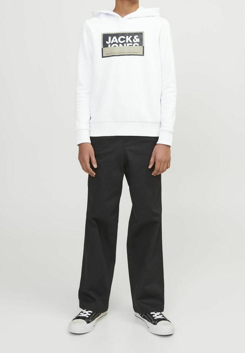 Personne portant un sweat à capuche blanc Jack & Jones, un pantalon large noir et des baskets noir et blanc, debout devant un fond clair uni.