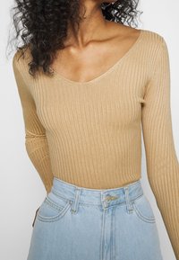 Kobieta nosząca beżowy prążkowany top z długim rękawem i dekoltem w serek, włożony w jasnoniebieskie dżinsy z wysokim stanem z guzikiem marki Lee.