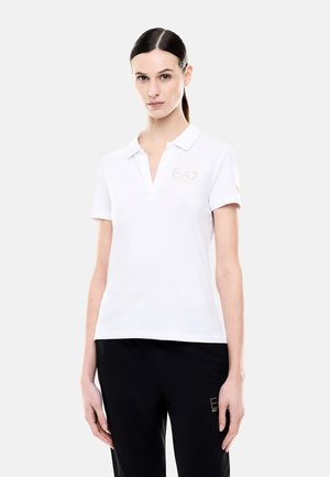 Donna con capelli scuri raccolti in una coda di cavallo indossa una polo bianca a maniche corte con logo EA7 e pantaloni neri con logo abbinato su sfondo semplice.