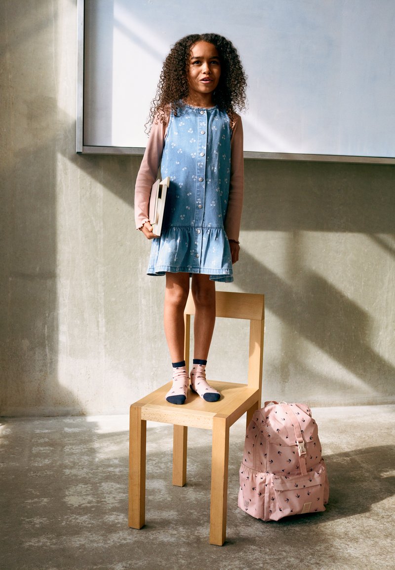 Νεαρό κορίτσι με φόρεμα από τζιν στέκεται ξυπόλητο σε ξύλινη καρέκλα κρατώντας βιβλία, δίπλα σε ροζ σακίδιο σε σκυρόδεμα δάπεδο.