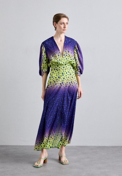 Maxi-jurk in een verlopen blauw en groen met een luipaardprint. Heeft een diepe V-hals, pofmouwen en een gestrikte taille. Vergezeld van groene sandalen.