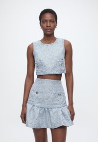 Γυναίκα που φοράει ανοιχτό μπλε cropped τοπ χωρίς μανίκια με υφή και αντίστοιχη φούστα με ψηλή μέση, εμπρός τσέπες και κουμπιά, στέκεται μπροστά σε λευκό φόντο.