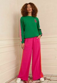 Sudadera verde con mangas rosas, que presenta un pequeño gráfico. Pantalones anchos rosas con rayas laterales naranjas. Zapatillas blancas.