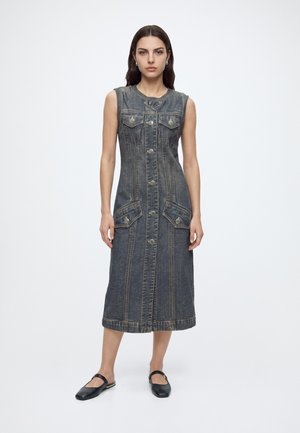 Femme debout portant une robe en jean sans manches, boutonnée, longueur genou, avec des poches avant et des chaussures plates noires sur fond blanc.
