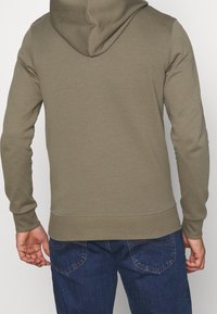 Sudadera con capucha de color verde oliva hecha de una tela suave y texturizada, con capucha ajustable con cordones, mangas largas y dobladillo acanalado, combinada con unos vaqueros azules.