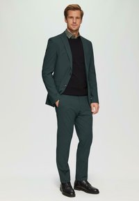 Dunkelgrüner Anzug bestehend aus einem maßgeschneiderten Blazer und Slim-Fit-Hosen, getragen über einem schwarzen Pullover mit einem Hemd darunter. Schwarze Schuhe.