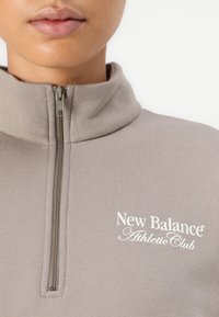 Heled hall zip-up sviiter kõrge kraega, millel on tekstureeritud kangas, väike tõmblukk ja valge "New Balance Athletic Club" logo.