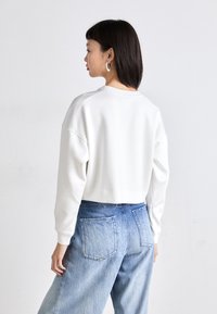 Κομμένο λευκό φούτερ με μακριά μανίκια, ραμμένες μανσέτες και στρογγυλή λαιμόκοψη, σε συνδυασμό με ανοιχτό μπλε, ξεθωριασμένο παντελόνι καμπάνα.