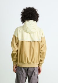 Hooded jacket with a beige upper section and a tan lower section, featuring elastic cuffs and a drawstring hem. Smooth, lightweight fabric.