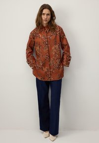 Cămașă de culoarea ruginii cu model floral, mâneci lungi, cu nasturi, asortată cu pantaloni evazați bleumarin și pantofi crem.