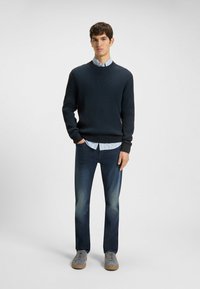 Marinblå ribbad tröja över en ljusblå randig skjorta, tillsammans med mörka jeans och grå sneakers. En enkel, avslappnad outfit med en klassisk passform.