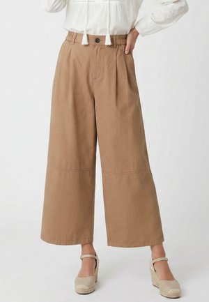 Beige weite, verkürzte Hosen mit Knopf und elastischem Bund, getragen mit hellbeigen Keilschuhen und einer weißen langärmeligen Bluse.