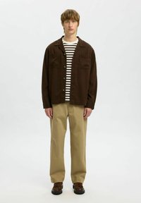 Brązowa kurtka zapinana na guziki na górze, noszona na pasiastą koszulę, beżowe spodnie i brązowe buty. Gładki materiał, luźny krój, dwie kieszenie na piersi w kurtce.