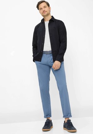 Uomo in piedi, indossa una camicia nera con bottoni sopra una maglia bianca, pantaloni azzurri chiari con cintura e scarpe da ginnastica scure con suole in gomma.