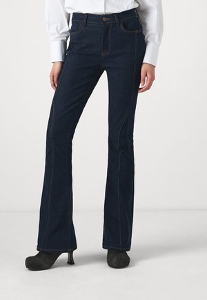 Jeans en denim bleu foncé évasés avec des coutures orange contrastantes, cinq poches et une fermeture à bouton. Le tissu semble lisse avec une légère texture.