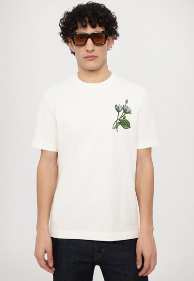 Weißes Baumwoll-T-Shirt mit kurzen Ärmeln, das ein floral gesticktes Design in Blau und Grün auf der linken Brust zeigt. Dazu getragen eine braun getönte Sonnenbrille.