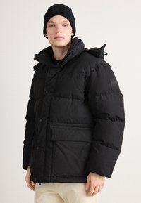 Black puffer jacket with a zip and snap closure, high collar, side pockets, and a logo patch on the sleeve. Worn with a black beanie.