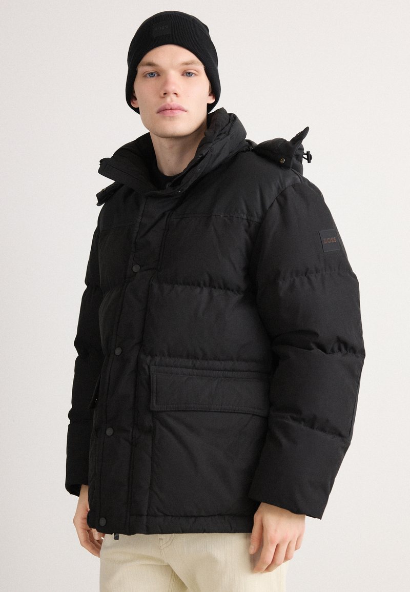 Black puffer jacket with a zip and snap closure, high collar, side pockets, and a logo patch on the sleeve. Worn with a black beanie.