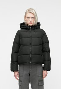 Jauns cilvēks ģērbjas melnā piepūstajā jakā ar kapuci un pelēk�ās kabatbiksēs, stāvot uz vienkārša gaiša fona.