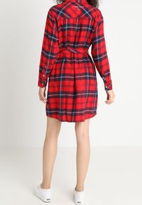 Abito camicia a quadri rossi e blu navy con maniche lunghe, vita cintrata e polsini con bottoni; presenta un dettaglio logo sul retro e arriva sopra il ginocchio.