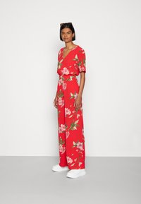 Macacão floral vermelho com mangas curtas, decote em V, cintura ajustada, pernas largas, apresentando flores cor-de-rosa e folhas verdes. Usado com ténis brancos.