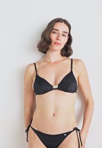 Juodas bikini komplektas, kuriame yra trikampio formos braletė su plonomis petnešėlėmis ir etikete, derinama su žemai sėsančiomis trumpikėmis, su raišteliais šonuose.