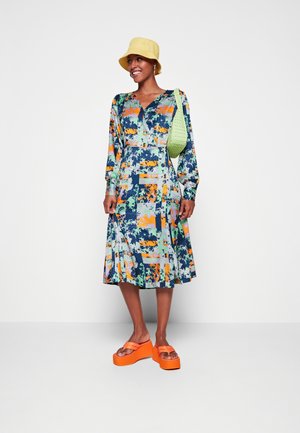 Vestido colorido e estampado com mangas longas, cintura ajustada e bainha larga. Combinado com sapatos de plataforma laranja brilhante e um chapéu amarelo.