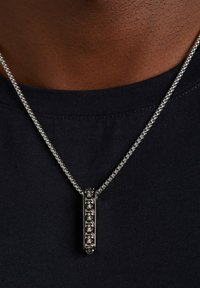 Zilveren ketting met een rechthoekige hanger met een gestructureerd ontwerp, die boven een donkere stof top hangt.
