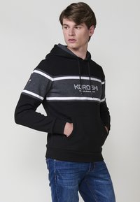 Un tânăr stă de profil, purtând un hanorac cu dungi negre și gri, având un logo, împreună cu o pereche de blugi, pe un fundal simplu.
