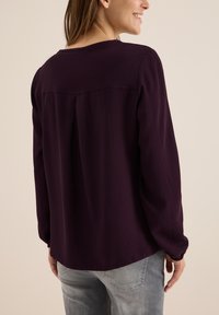 Blouse à manches longues en violet profond, présentant une texture côtelée subtile, un col rond et un détail plié à l'arrière. Assortie avec un jean clair.