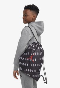 Crni ruksak svezan konopcima s ponovljenim bijelim natpisom "JORDAN" i crvenim Jumpman logom, napravljen od glatkog materijala, drži ga osoba u sivoj odjeći.