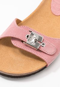 Nära bild av en rosa mockasandalrem med en glänsande silverspänne på en beige fotbädd och svart sula.