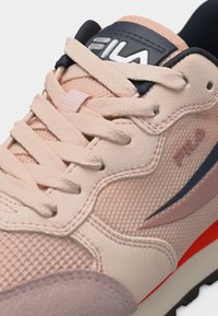 Κοντινό πλάνο ενός ροζ αθλητικού παπουτσιού FILA με μπεζ κορδόνια, υφασμάτινη υφή, δερμάτινες επιφάνειες, ναυτικό μπλε και πορτοκαλί λεπτομέρειες, και λογότυπο FILA στη γλώσσα και στη πλευρά.