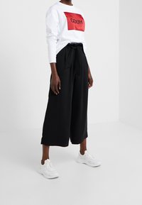Felpa bianca con logo rettangolare rosso, pantaloni neri a gamba larga con vita regolabile e scarpe da ginnastica bianche. Tessuto morbido, design casual.
