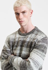 Grau-weißer Karo-Pullover aus einem strukturierten, fusseligen Stoff; mit einem runden Ausschnitt und langen Ärmeln, die unterschiedliche Farbtöne und Muster zeigen.