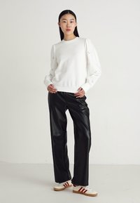 Sudadera blanca con puños acanalados, pantalones de piel sintética negra y zapatillas beige con rayas rojas. Tejido suave con ligero brillo.