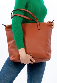 Brown textured leather tote bag with two black-accented handles, held by a person wearing a green ribbed sweater and jeans.
