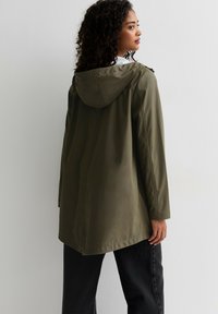 Olivgrön huvfodrad parka med en mjuk textur, som har en avslappnad passform, sidofickor och en knälång design. Bakifrån syns detaljer med sömmarna.