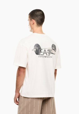 Mand iført hvid T-shirt med sort vægtstang og "EA7 Emporio Armani" logo på bagsiden, kombineret med beige shorts med logotryk.