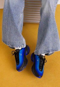 Le scarpe da ginnastica nere e blu presentano un design ondulato con una tomaia testurizzata, una suola intermedia prominente e lacci su uno sfondo contrastante di denim e giallo.