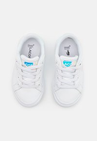 Baskets blanches avec une tige synthétique lisse, des lacets plats et une semelle en caoutchouc. Présente un logo Lacoste bleu et irisé sur la languette.