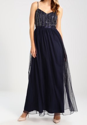 Kvinde iført en marineblå aftenkjole med en perlebesat overdel, tynde stropper og et flydende tylskørt, kombineret med nudefarvede spidse hæle.