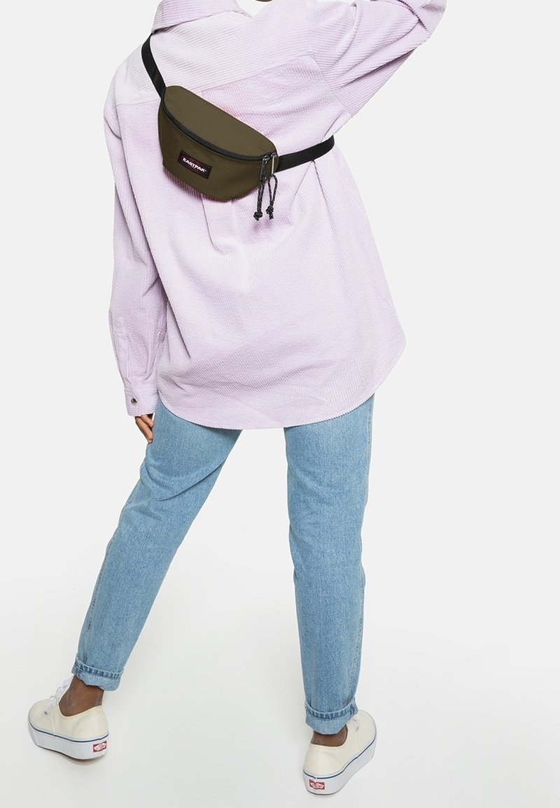 Personne portant une chemise violet clair, un jean bleu, des baskets blanches et une sacoche Eastpak vert olive portée en travers du dos.