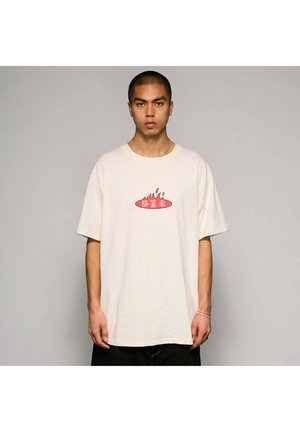 Młody mężczyzna stojący na jednolitym tle, ubrany w luźną białą koszulkę z czerwonym płomieniem i chińskimi znakami na piersi.