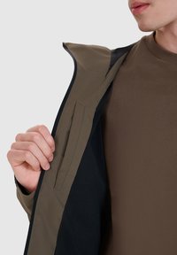 Personne portant un pull marron déboutonne une veste vert olive, révélant une poche intérieure et une doublure en mesh noire.