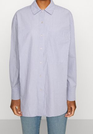 Persoon die een lichtblauw en wit verticaal gestreept overhemd met knopen en een zak draagt, gecombineerd met lichtgewassen blauwe spijkerbroek.