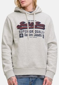 Lys grå hoodie med teksten "Jack & Jones Superior Quality Denim Goods" båret af person med tatoveringer på hals og hænder.